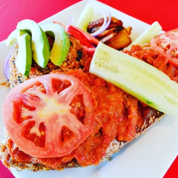 African dish with rice, avocado, plantains, and fish topped with tomato sauce and cucumber.