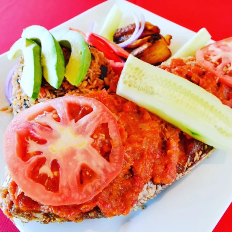 African dish with rice, avocado, plantains, and fish topped with tomato sauce and cucumber.