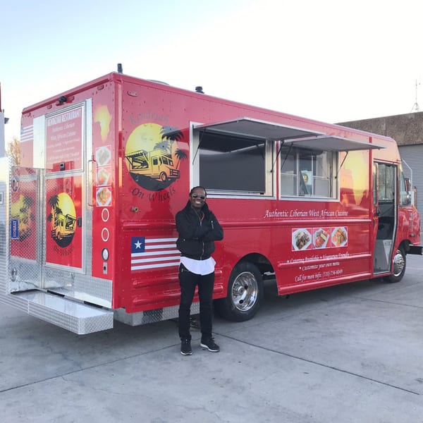 A man stands proudly beside the red Kendejah Restaurant food truck, featuring Liberian West African Cuisine.