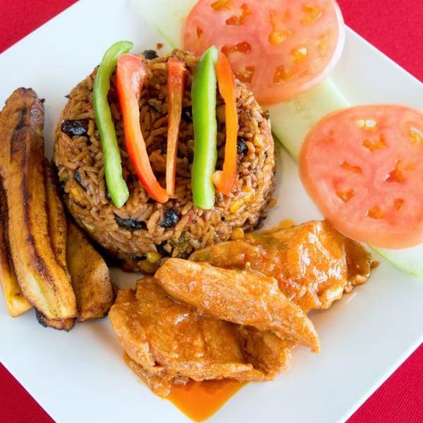 African dish with rice, plantains, chicken in sauce, and tomato and cucumber slices.