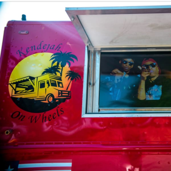 Two men in sunglasses look out from the serving window of a red Kendejah food truck.