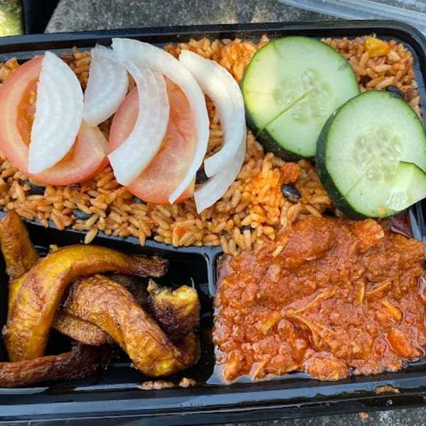 African meal with jollof rice, stewed meat, fried plantains, sliced tomatoes, onions, and cucumbers.