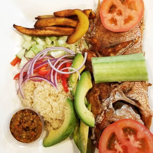 African fried fish with plantains, couscous, avocado, and a side of sauce on a white plate.